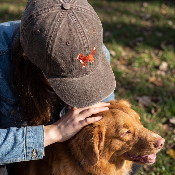 Person wearing a cap with a dog design, petting a brown dog outdoors.