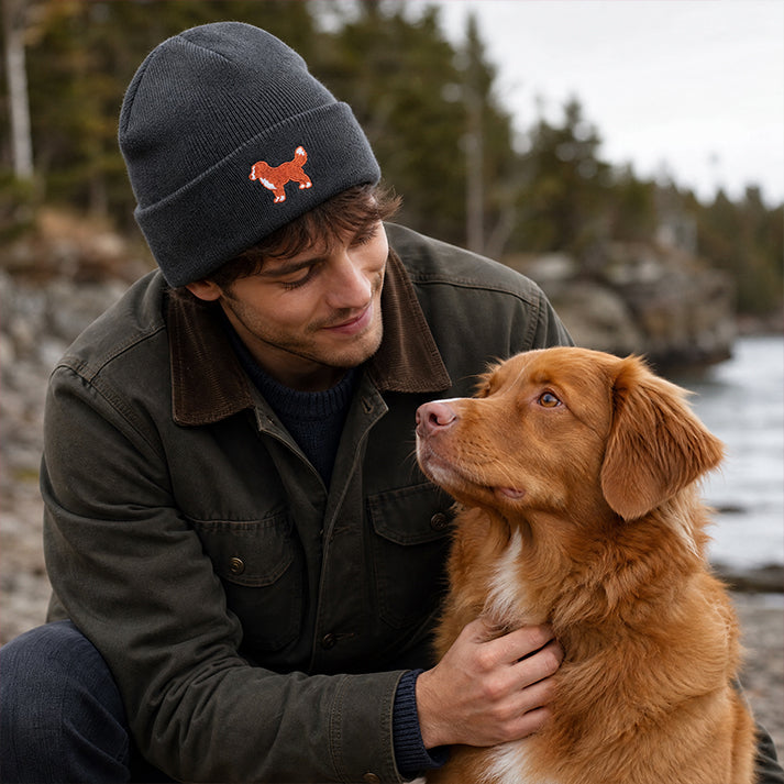 Man wearing a beanie with a dog logo, sitting with a brown dog by a body of water.