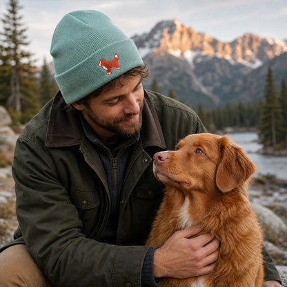 Man wearing a green beanie with a fox emblem, holding a brown dog in a mountainous landscape.