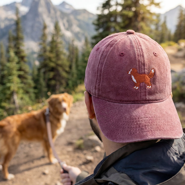 Person wearing a maroon cap with a dog logo, walking a dog on a trail with mountains and trees in the background.